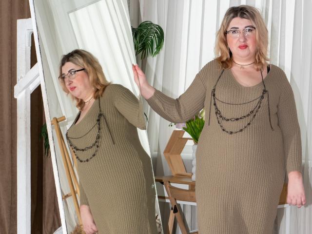 HelenEnigma, com cabelo loiro, óculos e um colar grosso, vestindo um vestido marrom texturizado, está em frente a um espelho, mostrando seu reflexo, segurando o espelho..