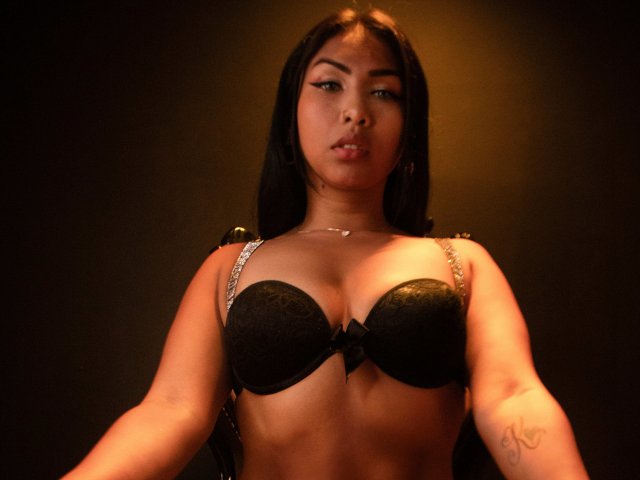 TracyLambo with dark hair, wearing a black bra, showing her tits, looking at the camera, subtle makeup, small tattoo on her right arm, warm lighting, neutral expression.