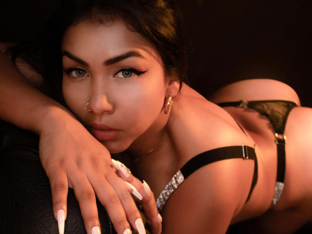 TracyLambo with dark hair, blue eyes, and a nose ring, wearing black lingerie and lace, leaning forward, showing her ass, with manicured nails, looking seductively at the camera.