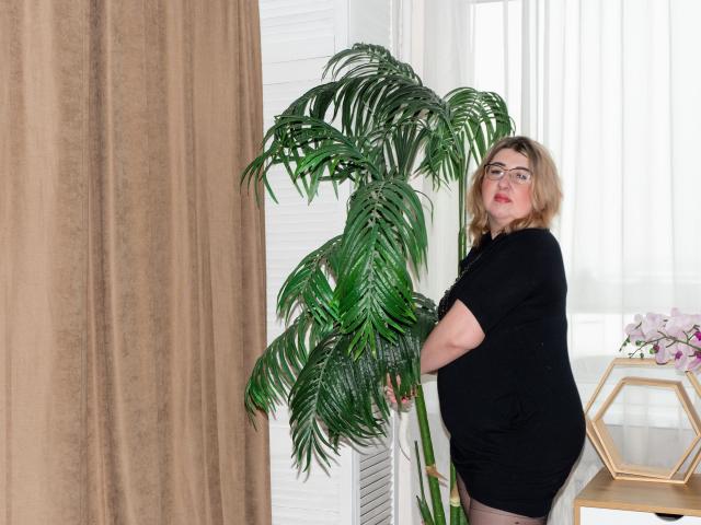 HelenEnigma, avec des lunettes, cheveux blonds, robe noire, debout à côté d'une plante verte, la tenant, regardant la caméra, les fesses légèrement appuyées contre la plante..
