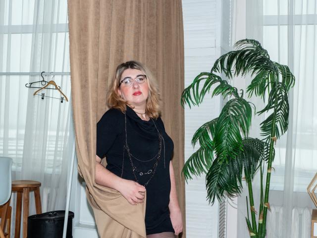 HelenEnigma, avec ses cheveux blonds, ses lunettes, son haut noir et son collier, tenant un rideau beige, debout à côté d'une plante verte en pot, avec des murs blancs et des cintres en arrière-plan. Ses fesses sont couvertes..