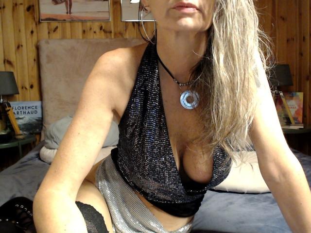 FrenchPlumeX with blonde hair, wearing a black mesh halter top showing her tits, silver skirt, and black thigh-highs, silver necklace, and hoop earrings, kneeling on a bed.