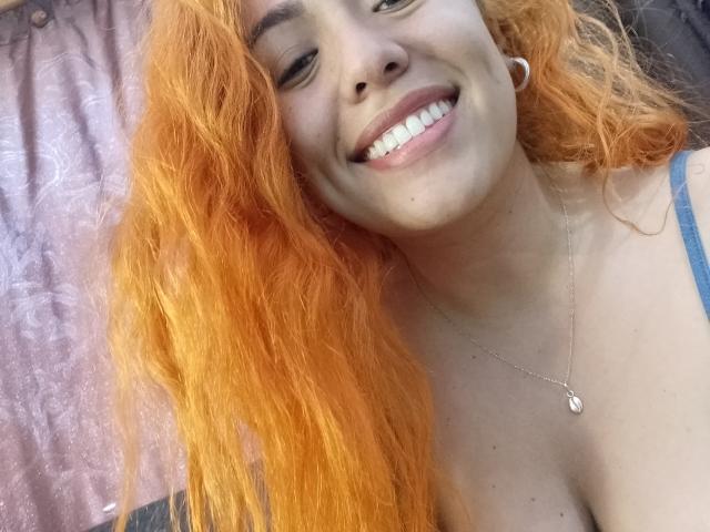 Joanallen met fel oranje haar, lachend, met haar witte tanden zichtbaar, draagt een blauwe band, een zilveren ketting en witte oordopjes, waarbij haar borsten gedeeltelijk zichtbaar zijn..