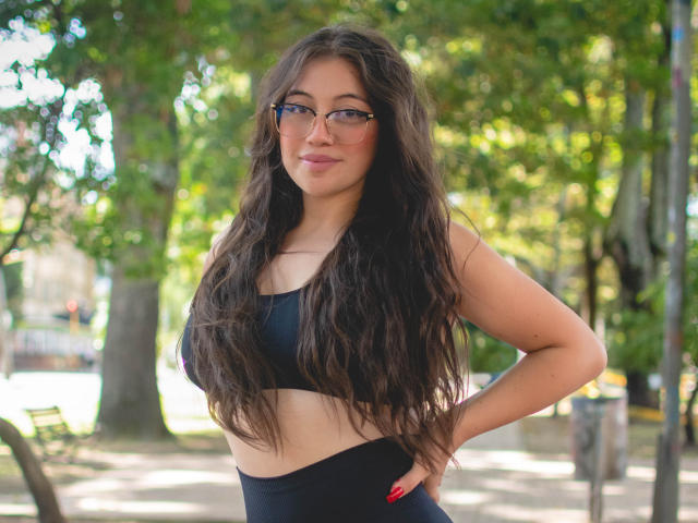 Amy Phillips mit langen, welligen braunen Haaren, die eine Brille trägt, ein schwarzes Sport-BH und schwarze, hochgeschnittene Hosen. Sie steht mit den Händen in den Hüften und lächelt leicht. Brüste sind sichtbar..