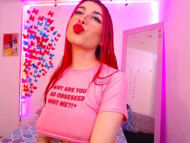 SammyViolet with red hair, pouting lips, wearing a pink shirt, large tits, in a pink-lit room, mirror reflecting her, colorful butterfly wall decoration.