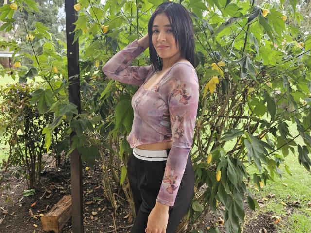 NicoColeman with medium tits, wearing a sheer pink floral top and black pants, standing in front of green foliage, touching her black hair with a slight smile.