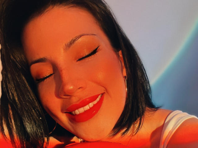 Liliysweet has red lipstick, closed eyes with winged eyeliner, and a smile. She has black shoulder-length hair, wearing a white top, and is holding her red-clad boobs.