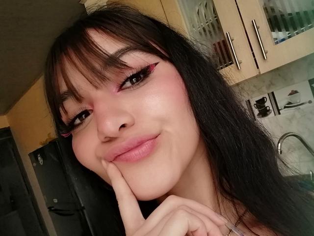 LindaWatsong has pink eyeliner, black hair with bangs, and is smiling with her finger on her chin, showing off her pink lips and fair skin. Her tits are not visible.
