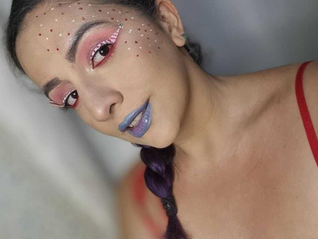 Andrea Sharick met paars gevlochten haar, rode eyeliner, roze oogschaduw, rode lipstick, blauwe lipstick op de onderlip, een rode top, een gezicht met stipjes en een gemiddelde borst, lachend..