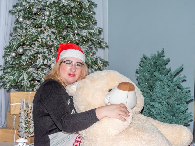 HelenEnigma met een bril, blond haar, een rode kerstmuts, een zwarte trui, die een grote witte teddybeer omhelst, lachend, staand voor een versierde kerstboom en een groene boom..