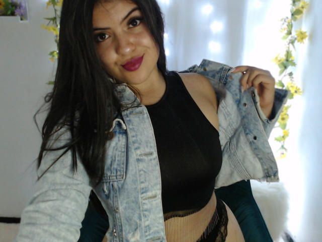 AlissonS, con cabello negro largo, lápiz labial rosa y una sonrisa, vestida con una chaqueta vaquera sobre un top corto negro y una falda beige de punto, mostrando sus pechos..