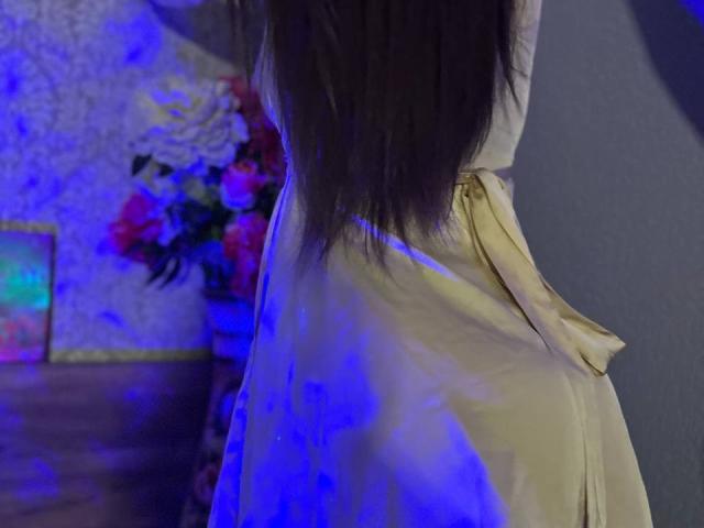 CheriLady is terug, met lang bruin haar, in een glanzende gele jurk met een spleet aan de zijkant, onder blauw licht. Haar achterkant is licht zichtbaar. Een bloemenboeket in de achtergrond..
