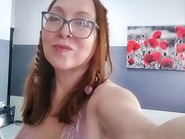 IrunchaArt has red-brown hair in braids, wearing glasses, and a pink lace top, showing a smile with a cute nose ring. Boobs visible in the upper frame.