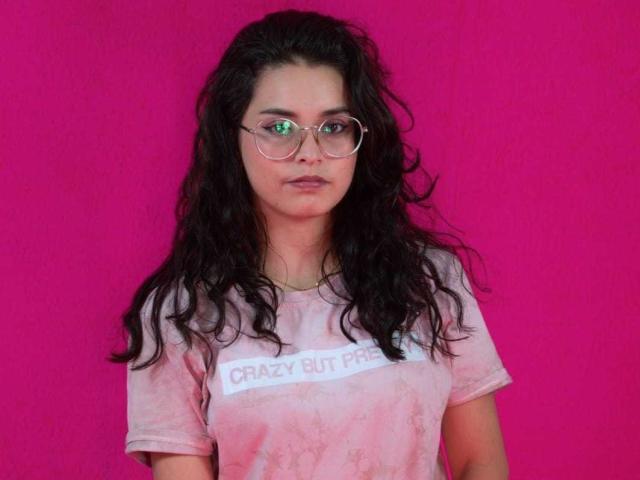 AhislinSoul with long, wavy black hair, wearing round glasses and a pink tie-dye shirt, standing against a bright pink background. She's got a serious, almost bored expression.