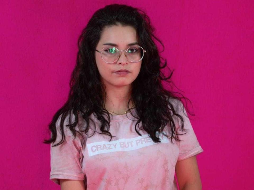 AhislinSoul with long, wavy black hair, wearing round glasses and a pink tie-dye shirt, standing against a bright pink background. She's got a serious, almost bored expression.