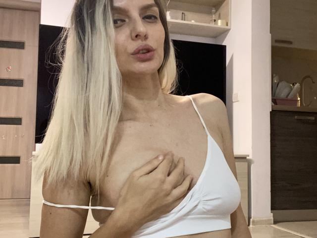 DouceAmelia with blonde hair, white top, and visible tits, covering one breast with her hand, in a modern indoor setting. She has pouty lips and a neutral expression.