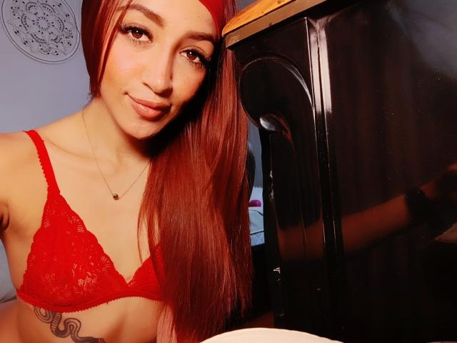 AnnaKhalo con un sujetador rojo, mostrando pequeños senos, cabello rojo largo, un tatuaje discreto, sonriendo, un armario oscuro al fondo, luz cálida, maquillaje sutil, collar, selfie casual..