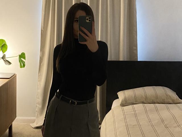 Dionisia with long dark hair, black turtleneck, gray high-waisted skirt, black belt, taking mirror selfie, showing her small tits, standing in dimly lit bedroom with bed.