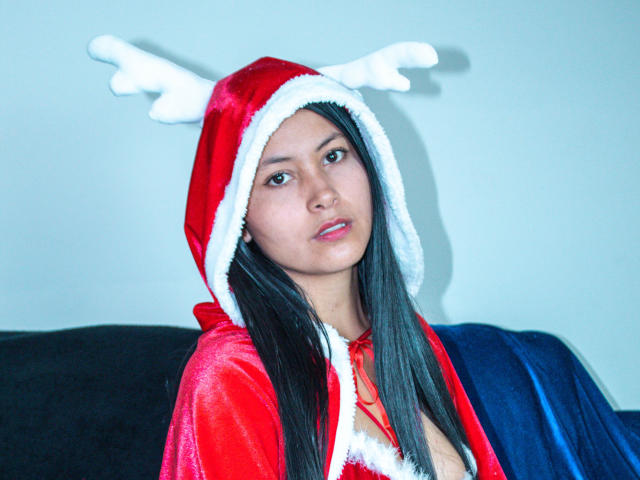 Emily Savage trägt ein rotes Weihnachtsmannkostüm mit weißem Pelzkragen, einen Stirnband mit Geweihen und hat langes, schwarzes Haar. Sie schaut neutral in die Kamera und zeigt kleine Brüste..
