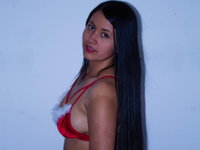 EmilySavage with long black hair, red velvet bra with white fur, small tits, looking over shoulder, light blue background, subtle smile, slight blush, red lipstick, medium skin tone.