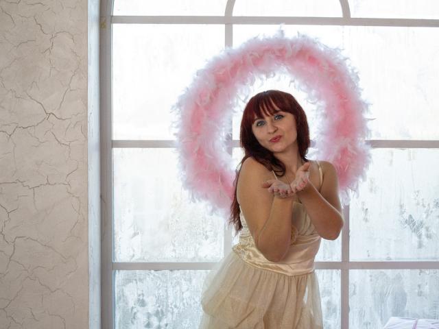 NinaFlawlees with red hair, wearing a pink and white feather halo, and a white lace dress, standing in front of a window, hands near her tits, smiling.