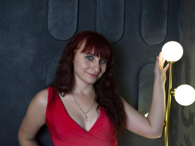 NinaFlawlees tem cabelo ruivo, olhos azuis e um sorriso adorável, vestindo um vestido vermelho com um decote baixo, que mostra seus seios, e segurando uma lâmpada com orbes brilhantes..