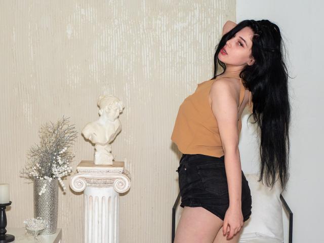 LiyaFrench, con cabello negro largo, un top de tirantes color piel y pantalones cortos negros, está de pie con un brazo detrás de la cabeza, mostrando sus glúteos pequeños y su figura delgada, junto a una estatua blanca sobre una columna..