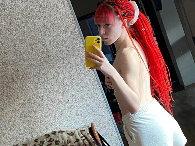 Topless PassionSupply with bright red braids and horns, holding a yellow phone, taking a mirror selfie. She's wearing white pants, showing her ass. Reflects in a textured wall.