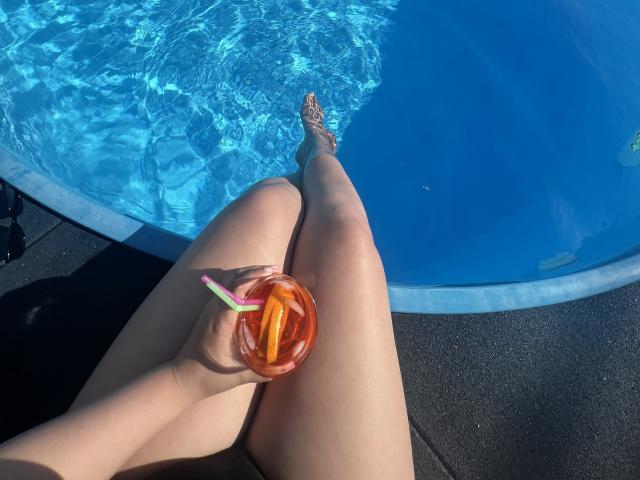 Barbarica's legs in a pool, holding a drink with an orange straw, her toes in the water, sunlit skin, black pool deck, clear blue water, relaxed, summer vibe.