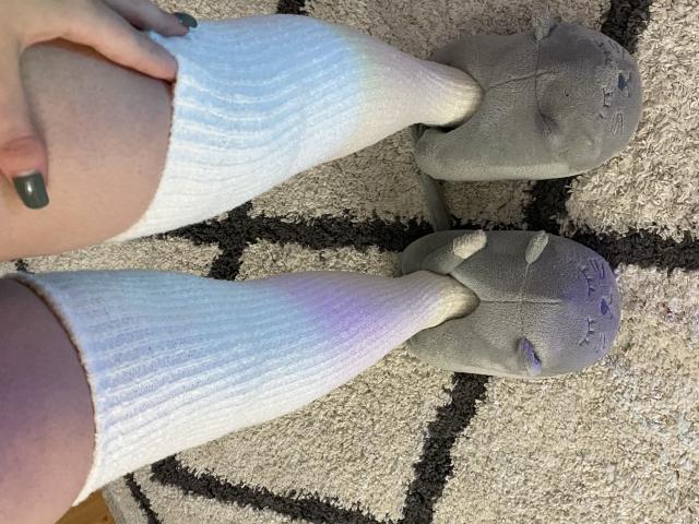 Rubbi's legs in white ribbed socks, gray slippers with visible wear, and green-painted nails, standing on a beige and black patterned carpet.