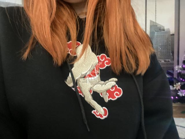 Le sweat à capuche noir de Rubbi, avec un motif de cheval argenté et rouge, dévoile légèrement ses seins. Ses cheveux orange sont visibles dans le cadre, avec un arrière-plan de paysage urbain flou..
