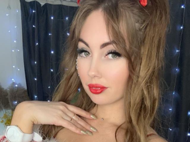 NinaTender with wavy brown hair, red lipstick, green eyes, long eyelashes, and manicured nails, touching her neck, wearing red bows in her hair, and blue fairy lights.