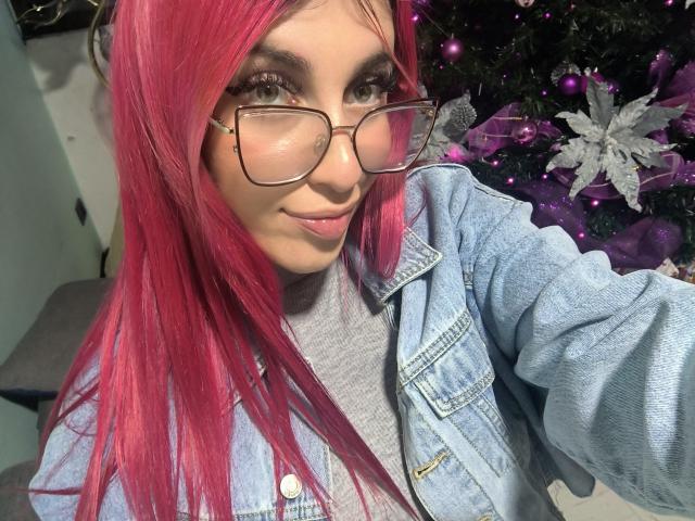 SammyViolet, con cabello rosa, gafas y una chaqueta vaquera, sonríe a la cámara, mostrando su linda cara y su pequeño nariz. Sus pechos están cubiertos por su camisa..