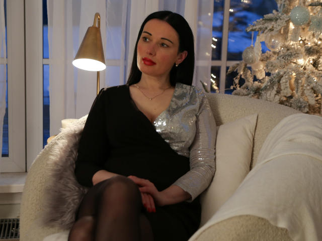 HottAlina met zwart haar, rode lippenstift, een zwarte jurk, een zilveren mouw met pailletten, zwarte leggings, zittend op een witte bank, een gouden lamp, een kerstboom met witte versieringen, een blauwe nacht buiten..