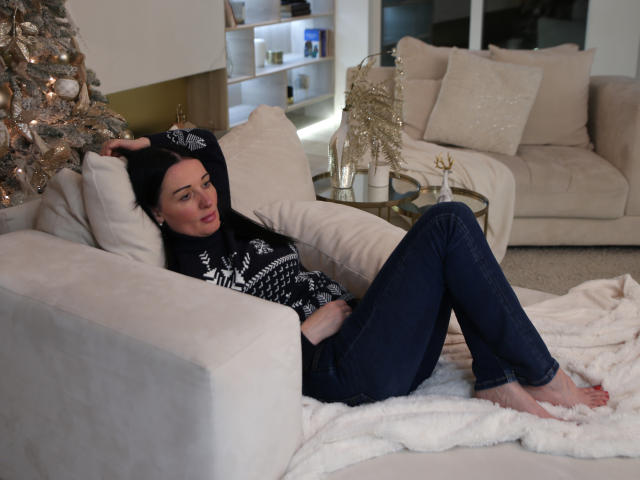HottAlina met bleke huid, een zwarte muts, een zwart-witte trui, blauwe jeans, blote voeten, liggend op een witte bank, armen boven het hoofd, ontspannen, naast een zilveren kerstboom, gezellige kamer..