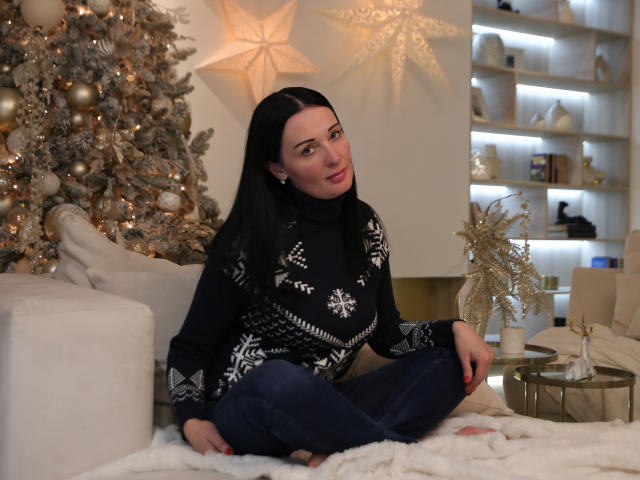 HottAlina, avec ses longs cheveux noirs, portant un pull noir orné de motifs blancs et un jean bleu, est assise en tailleur sur un tapis blanc, devant un sapin de Noël décoré..