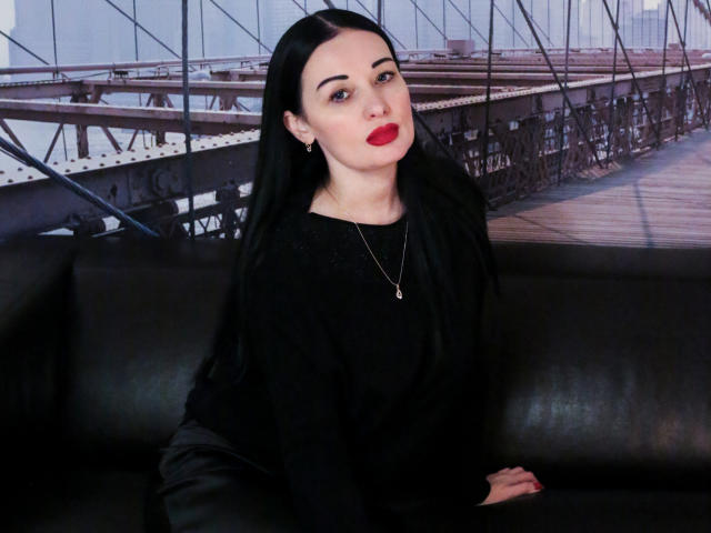 HottAlina, avec de longs cheveux noirs, du rouge à lèvres rouge, un pull noir et un collier, assise sur un canapé en cuir noir, regardant la caméra avec un léger sourire. Les seins ne sont pas visibles..