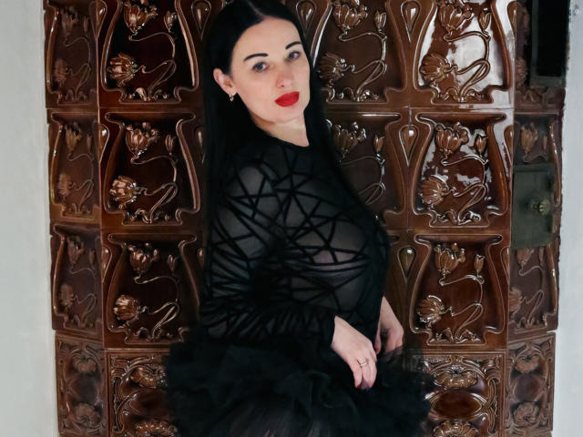 HottAlina com cabelo preto, batom vermelho e um vestido preto transparente que mostra seus seios, e um tecido preto estampado, apoiada em uma parede de azulejos marrons ornamentados..