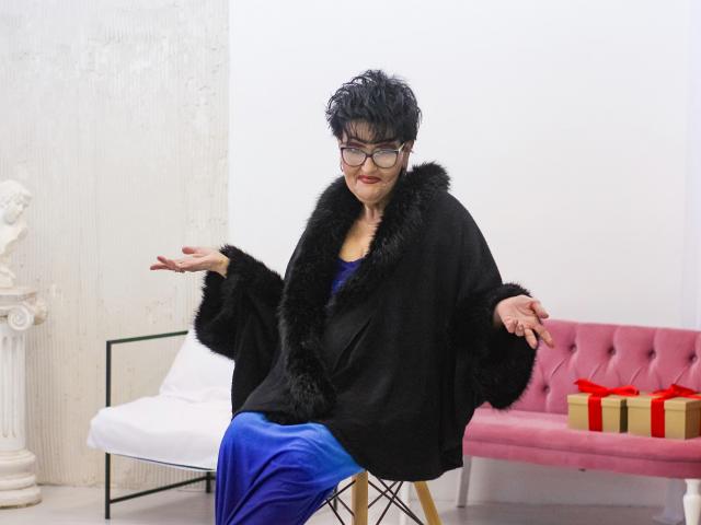 SondraRiddle with short black hair, glasses, black fur coat, blue dress, sitting on chair, arms spread, pink tufted sofa with red gift, white wall, classical statue.