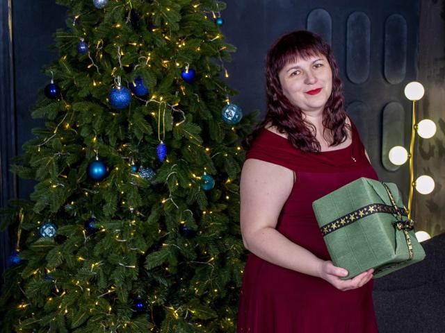 AliceInks with red dress, holding green gift, standing next to Christmas tree with blue ornaments and lights. She's got curly brown hair, red lipstick, and a cute smile.