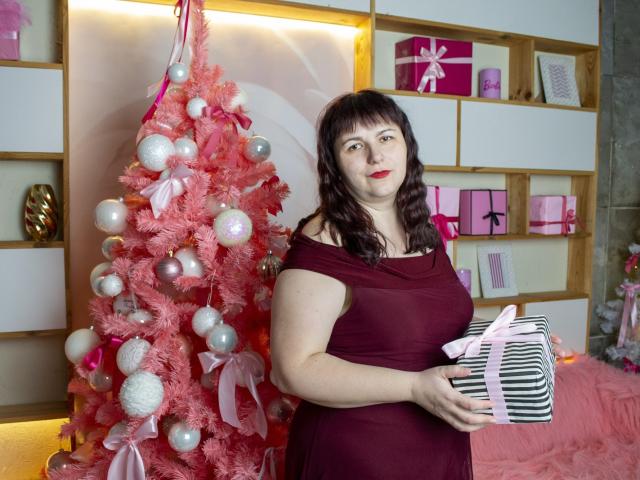 AliceInks with pale skin and red lipstick, wearing a maroon dress, holding a striped gift, standing next to a pink Christmas tree with white and pink ornaments.
