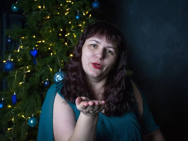 Alice, con i capelli castano ondulati, rossetto rosso, vestito color turchese e mano alzata, è in piedi di fronte a un albero di Natale con luci gialle e ornamenti blu..