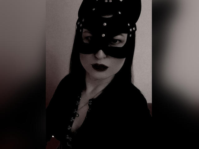 EvaBeautyAh, com batom escuro, máscara preta e um adorno preto na cabeça, exibindo o decote em uma blusa preta. Seus olhos estão semicerrados e ela parece sedutora. Imagem em tons de preto e branco, com sombras..