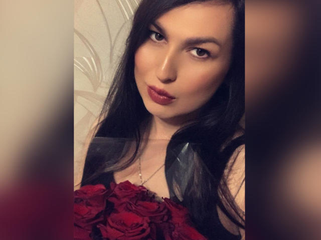 EvaBeautyAh with long black hair, red rose bouquet, glossy pink lips, and a slight pout, wearing a black top, in a soft, warm-lit, blurry photo.