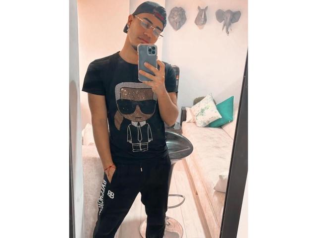 Jaden69 steht vor einem Spiegel und macht ein Selfie mit seinem Handy. Er trägt ein schwarzes T-Shirt mit einem Aufdruck, schwarze Hosen und eine Mütze, die verkehrt herum getragen wird, und eine Brille..