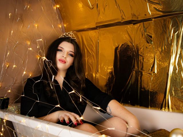 PamDares, con cabello castaño largo, lápiz labial rojo, vestido negro y una tiara dorada, sentada en una bañera, rodeada de luces de hadas, con su mano sobre su vulva desnuda..