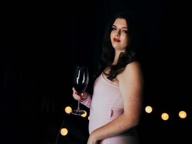 PamDares, avec ses cheveux noirs et ondulés, sa robe rose pâle, tenant un verre de vin rouge, sur un fond noir, avec des guirlandes lumineuses. Elle a un regard sexy et confiant, avec du rouge à lèvres..