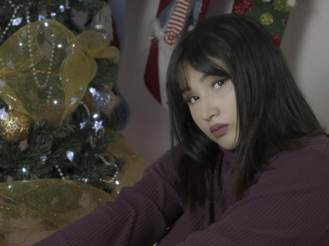 Valerie Collinss, avec ses cheveux bruns foncés, son pull violet, regardant la caméra, se tient à côté d'un sapin de Noël décoré avec un ruban jaune et des guirlandes blanches..