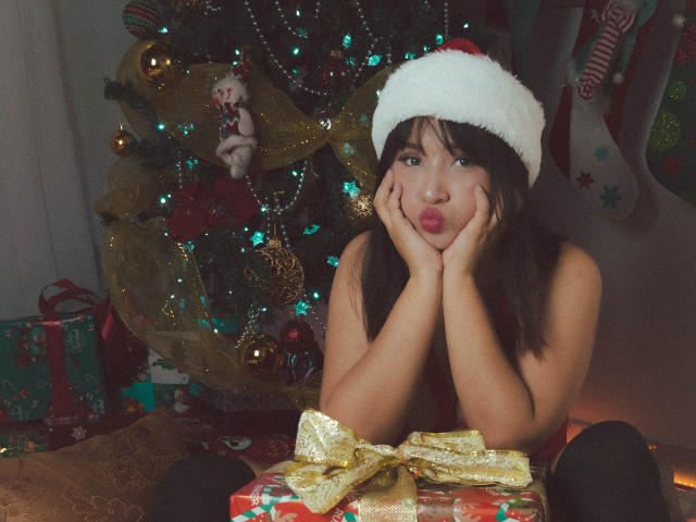 ValerieCollinss with pouty lips, hands on cheeks, wearing a Santa hat, sitting in front of a Christmas tree, hands on her face, and a golden wrapped gift in front.