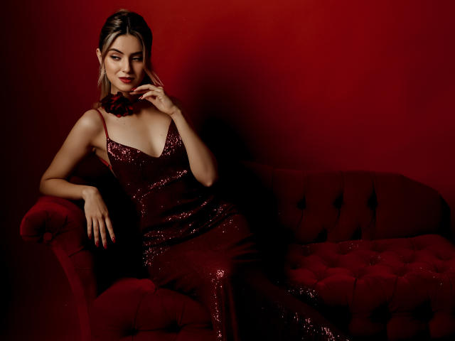 JasmineDay in a shiny red sequin dress, sitting on a red velvet couch, hand near mouth, showing off her tits and ass, with a red background.